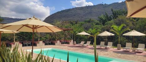 Zona de piscina con sillas de descanso y sombrillas, rodeada de vegetación y un paisaje montañoso.