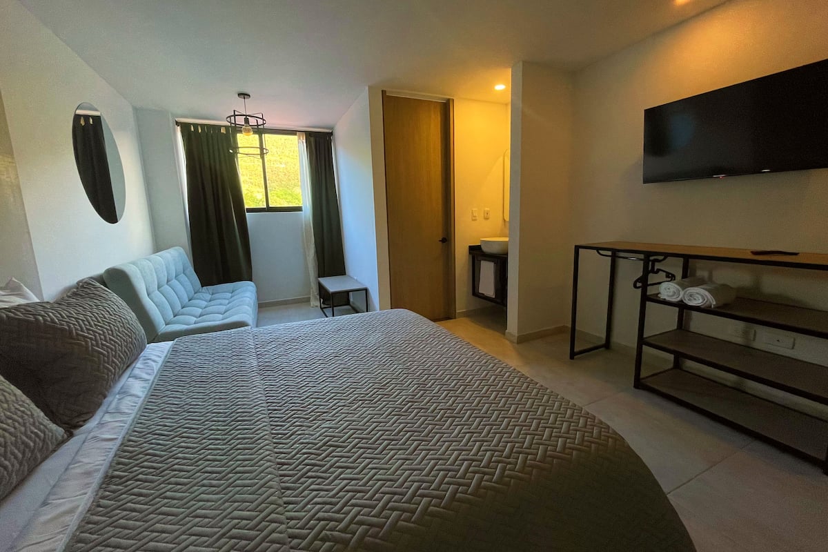 A hotel room with a large bed, a sofa, a small table, a TV, and a bathroom visible through an open door.