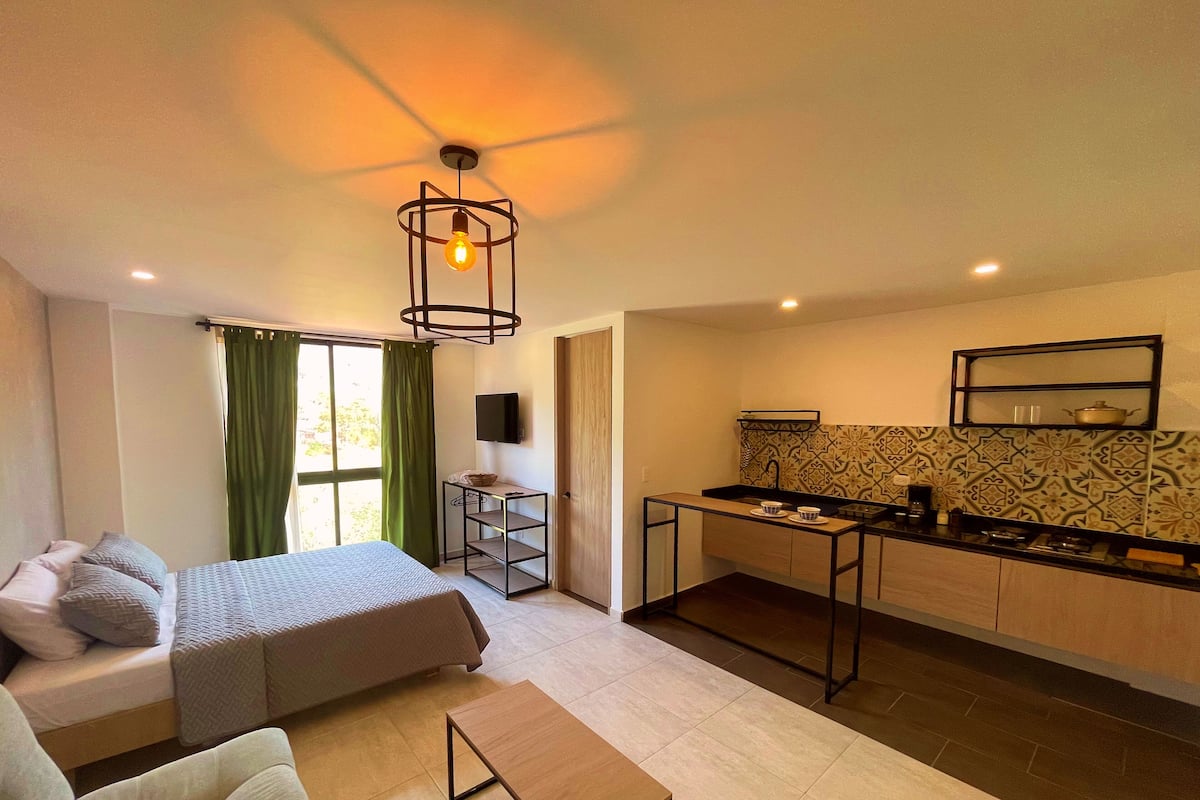 A compact hotel room with a bed, a sofa, a small table, a kitchenette, and a TV mounted on the wall.