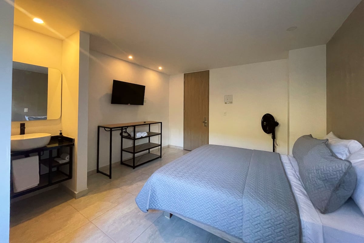 A modern bedroom with a bed, a bedside table, a TV, and a bathroom with a sink and mirror.