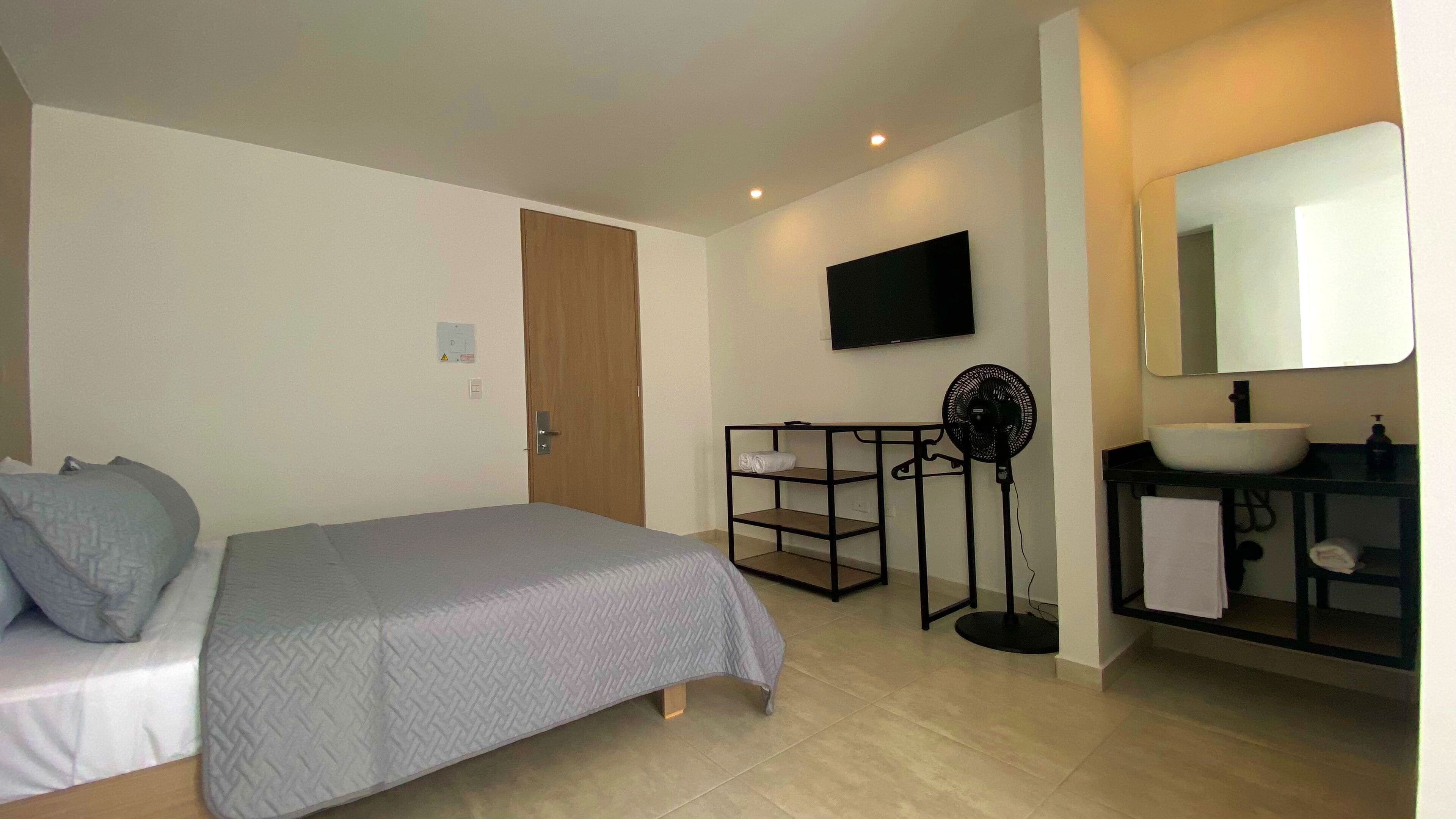 Una habitación de hotel con cama, televisor montado en la pared, ventilador y lavamanos con espejo.