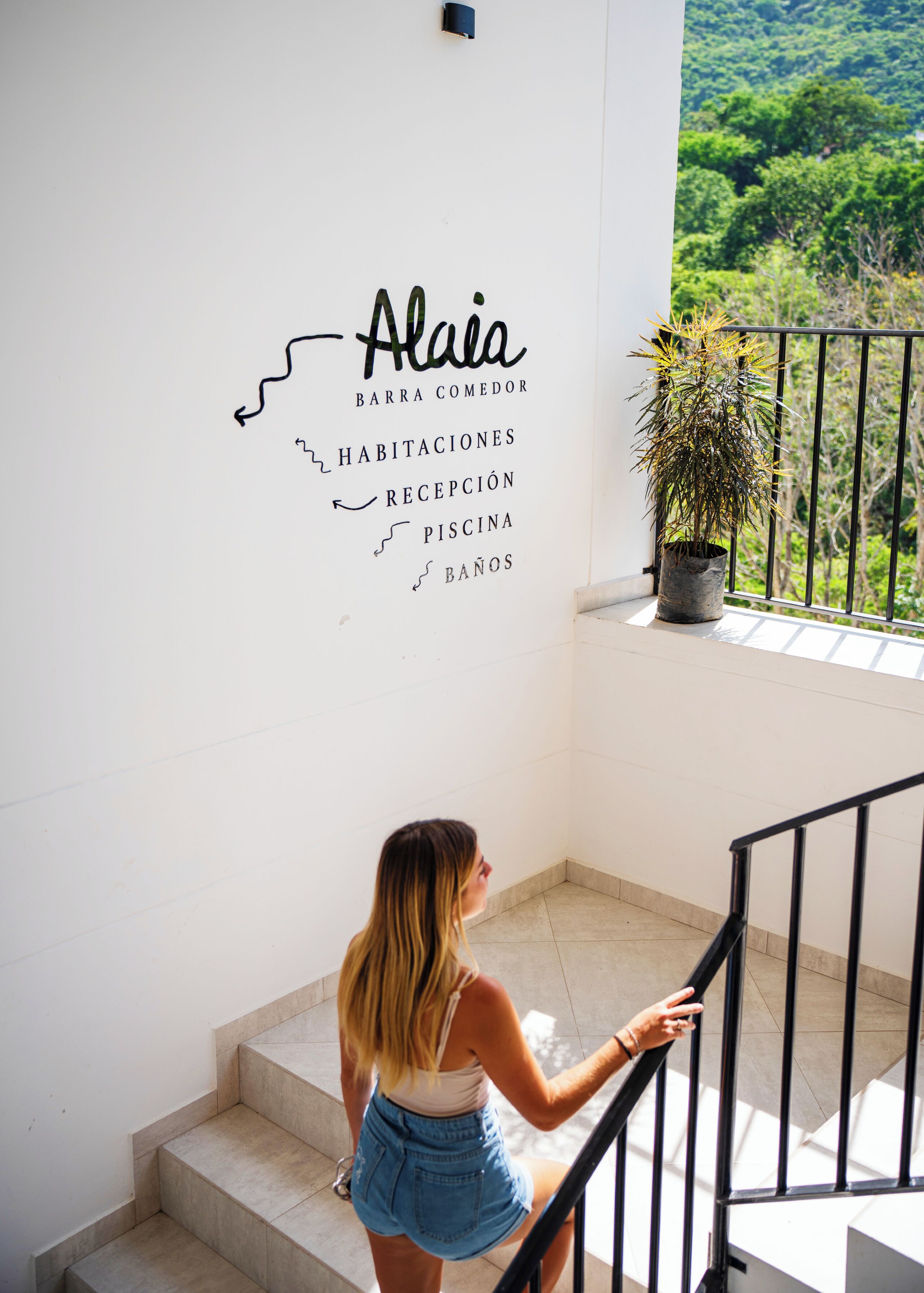 Una mujer bajando una escalera con un cartel que indica servicios como bar, comedor, habitaciones, recepción, piscina y baños.
