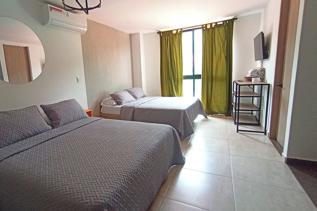 A hotel room with two beds, a round mirror, an air conditioning unit, a small table with a chair, and a TV mounted on the wall.