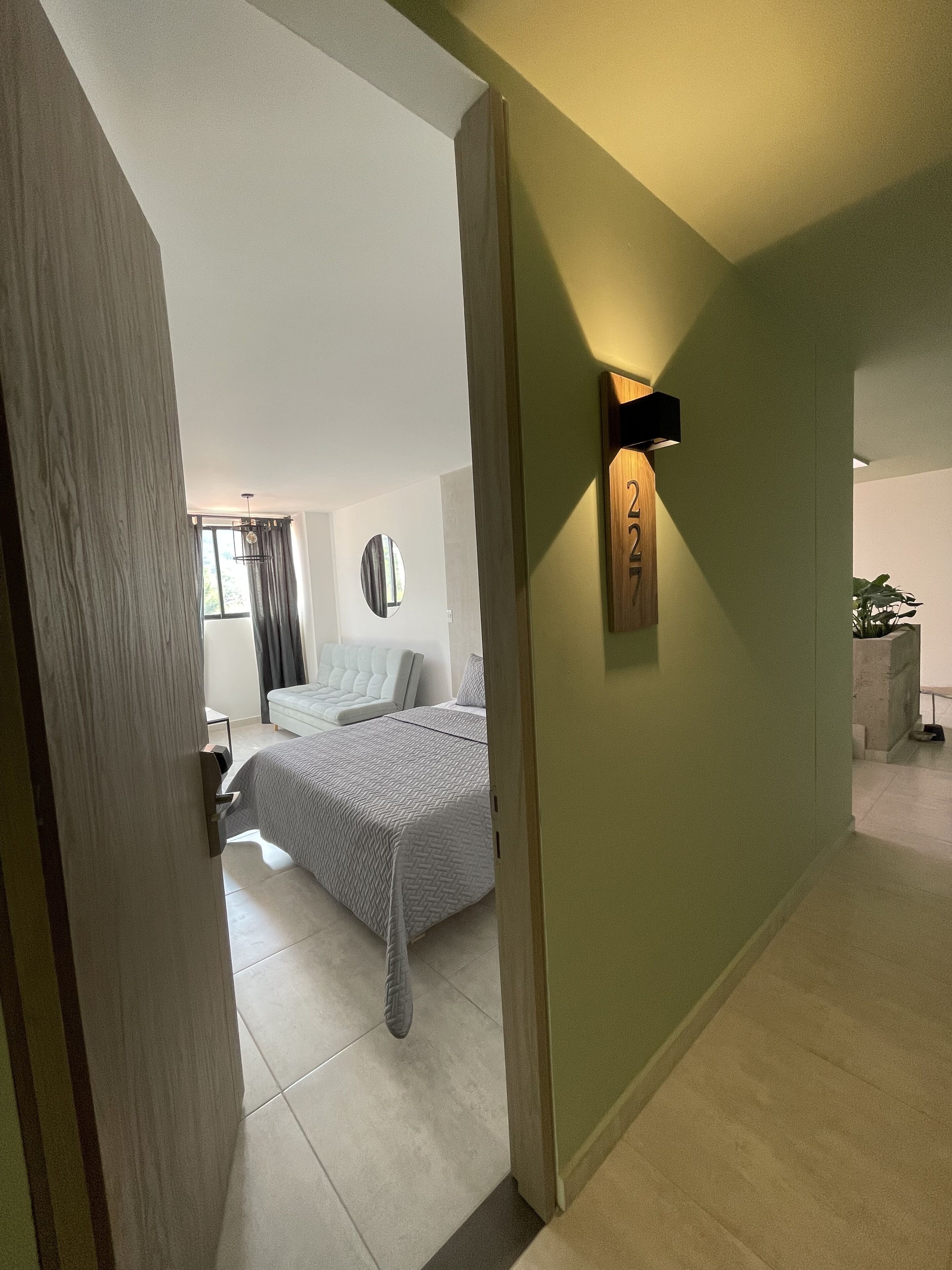 A modern hotel room with a bed, a bedside table, a mirror, and a wall-mounted light fixture.