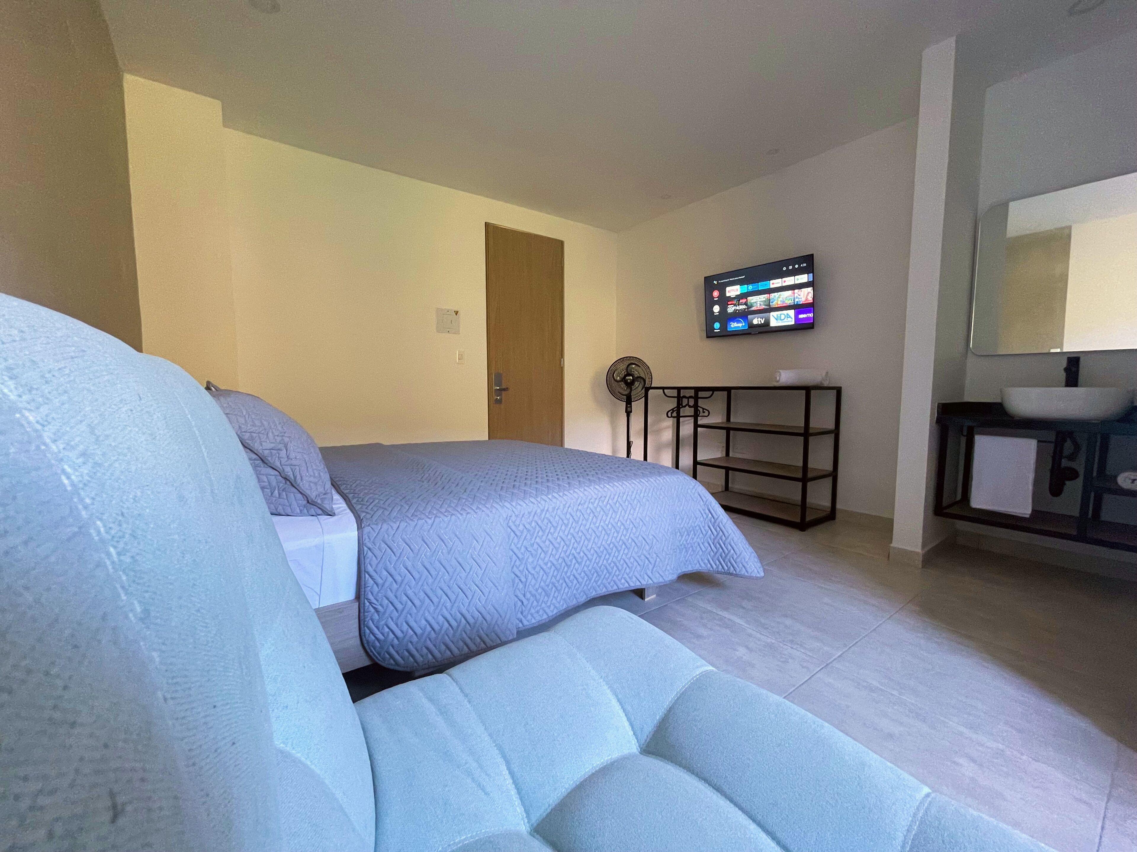 A bedroom with a bed, a bedside table, a TV mounted on the wall, and a bathroom with a sink and mirror.