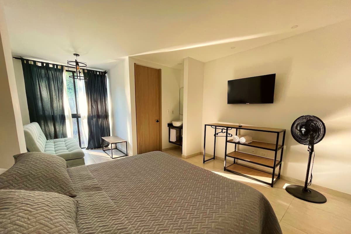 A hotel room with a large bed, a chair, a small table, a TV, and a standing fan.