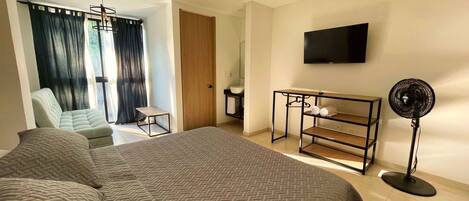 A hotel room with a large bed, a chair, a small table, a TV, and a standing fan.
