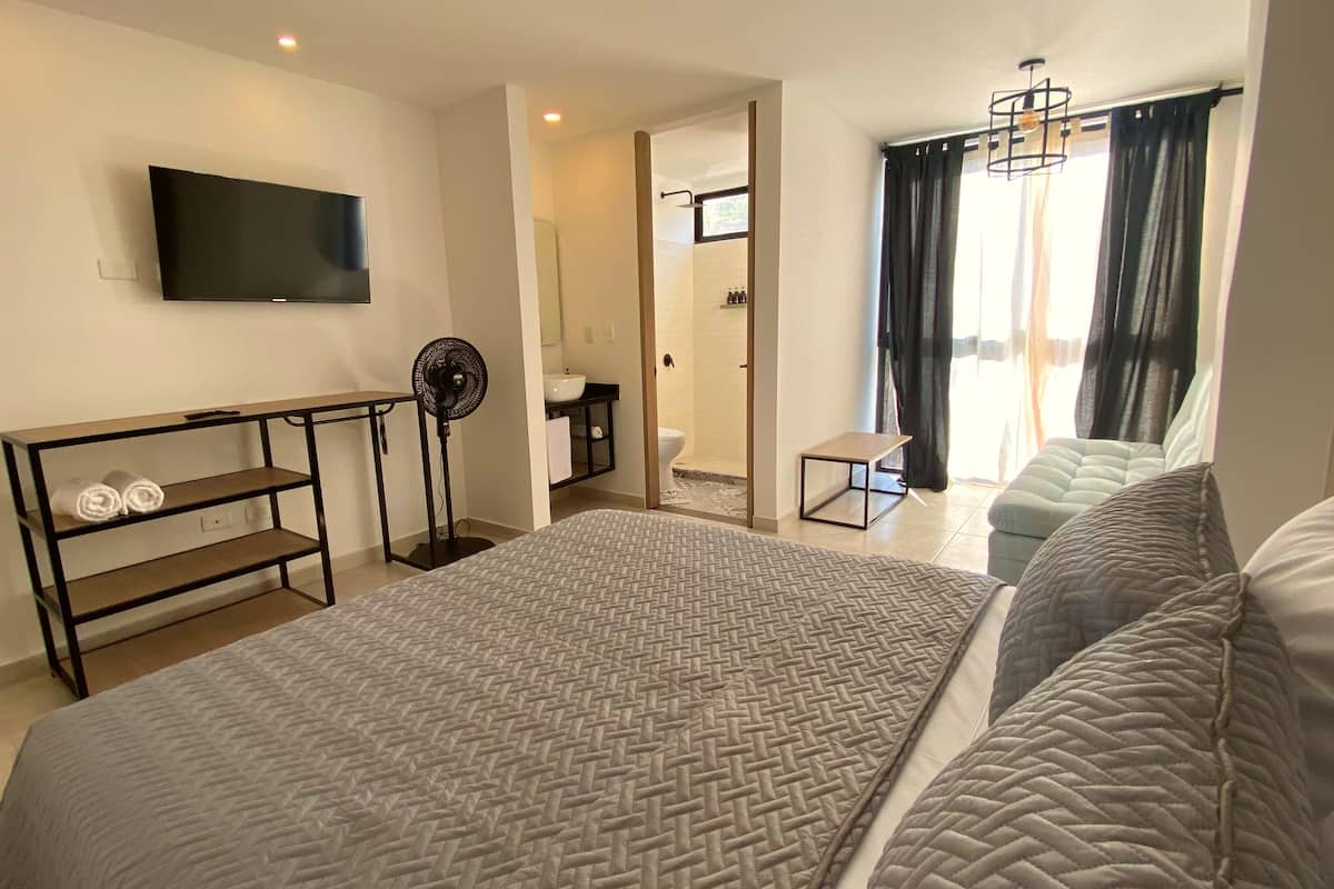 A hotel room with a large bed, a TV mounted on the wall, a desk with a fan, and a bathroom visible through an open door.