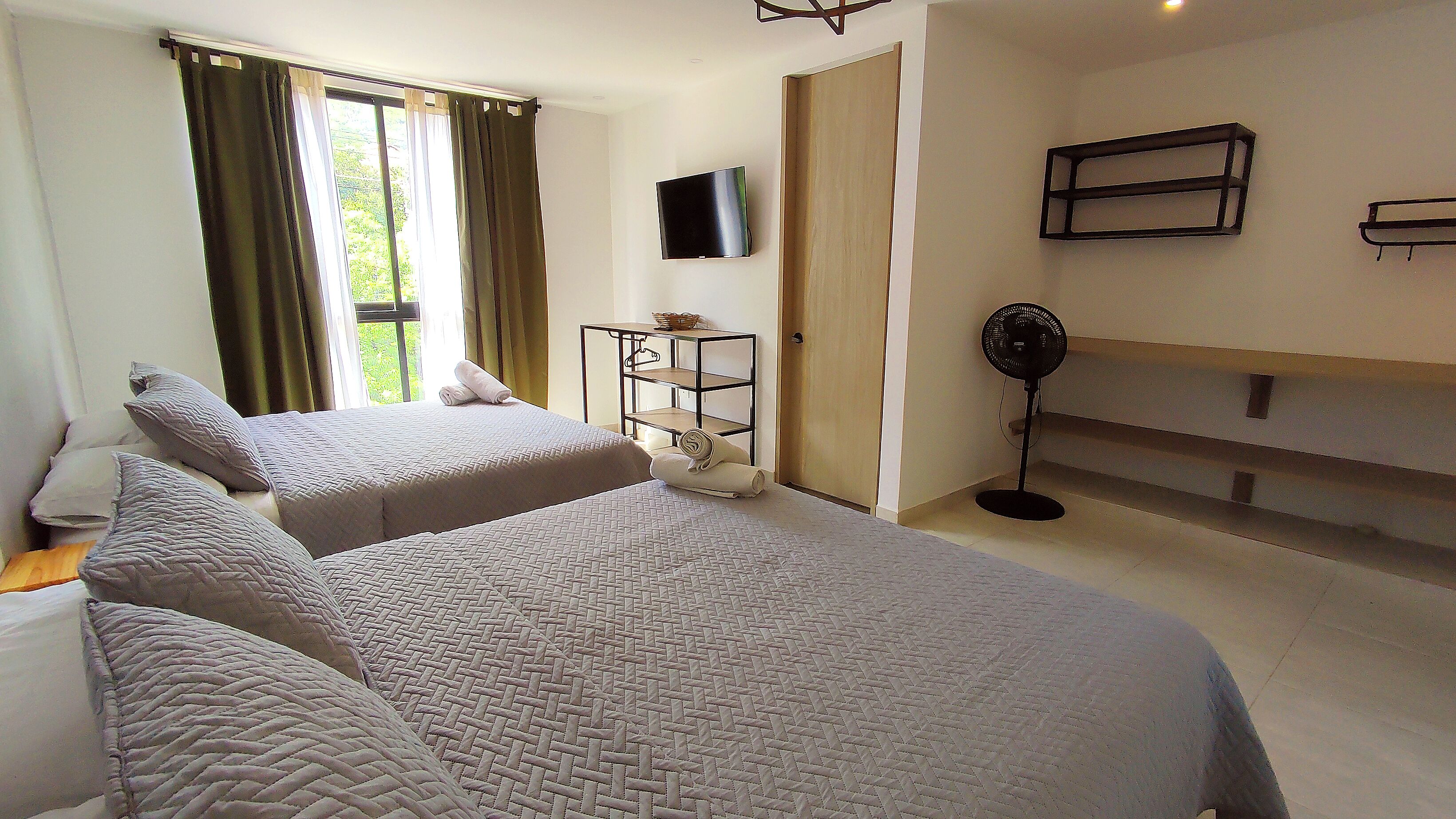A hotel room with two beds, a TV mounted on the wall, a standing fan, and a small shelf unit.