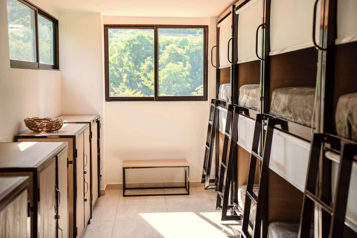A compact room with bunk beds, a small bench, a window with a view of greenery, and a cabinet with a basket on top.