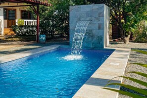Ein Pool mit Wasserfall, umgeben von einem Garten und einem Gebäude mit einer überdachten Terrasse.