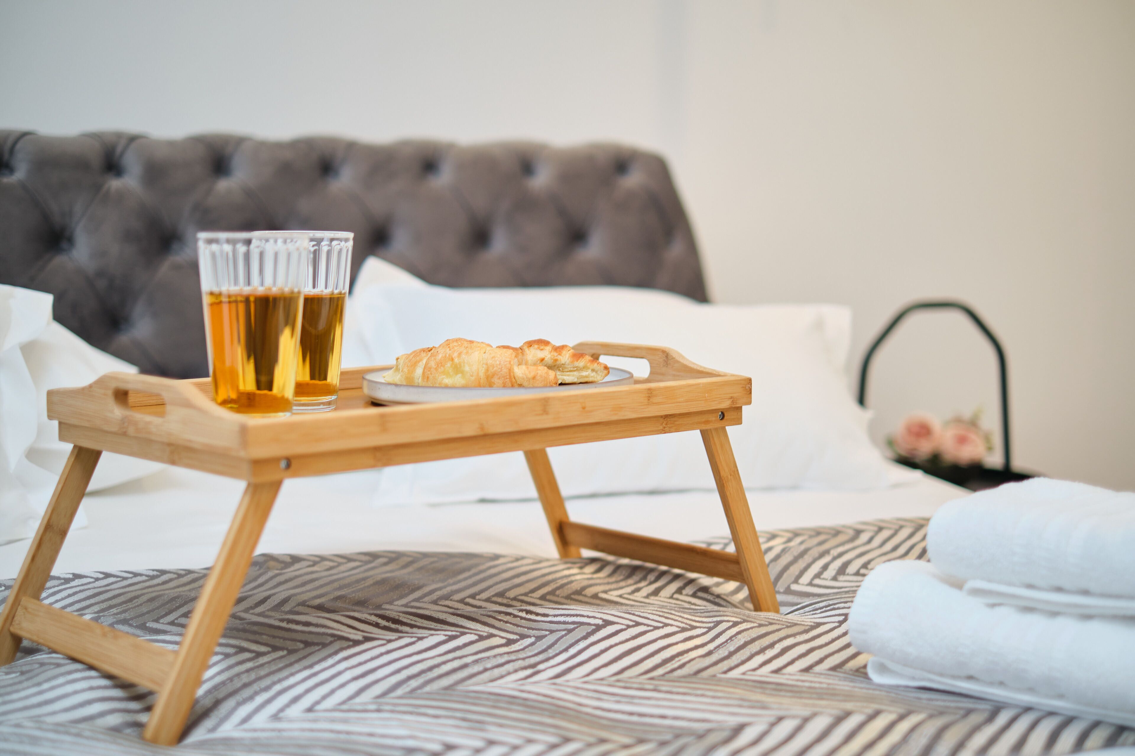 A bed with a tufted headboard, a wooden tray with a croissant and two glasses of beer, and a patterned bedspread.