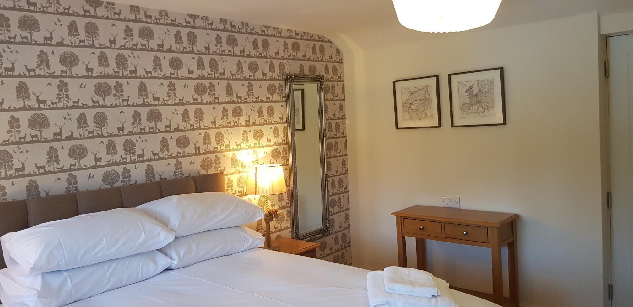 A hotel room with a bed, a nightstand with a lamp, a wooden dresser, and two framed pictures on the wall.
