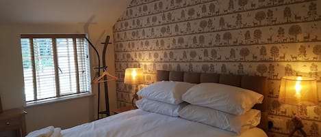 Una cama bien hecha con sábanas blancas, dos lámparas de noche, un perchero de madera y una ventana con persianas.