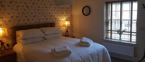 A neatly made bed with white linens and two folded towels on the side, a wall clock, and a window with blinds.
