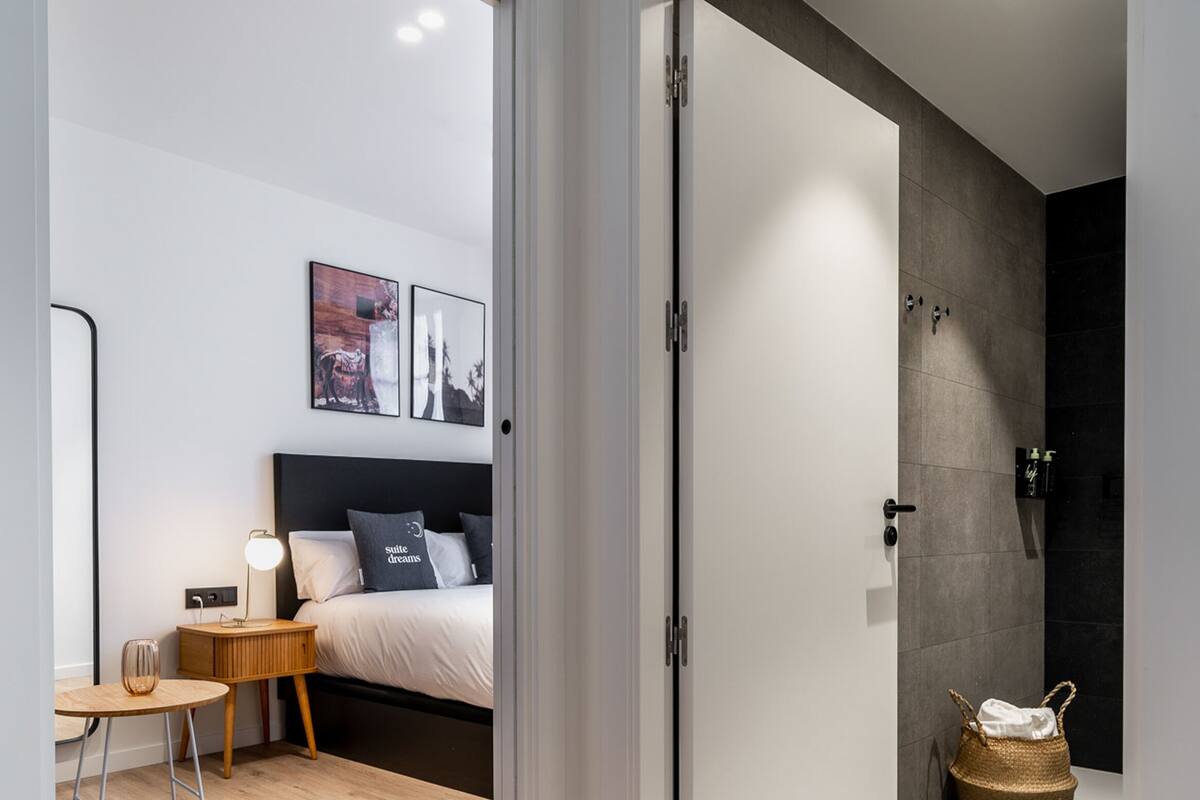 A modern bedroom with a wooden floor, a bed with white bedding, a bedside table with a lamp, and a wall-mounted shelf.