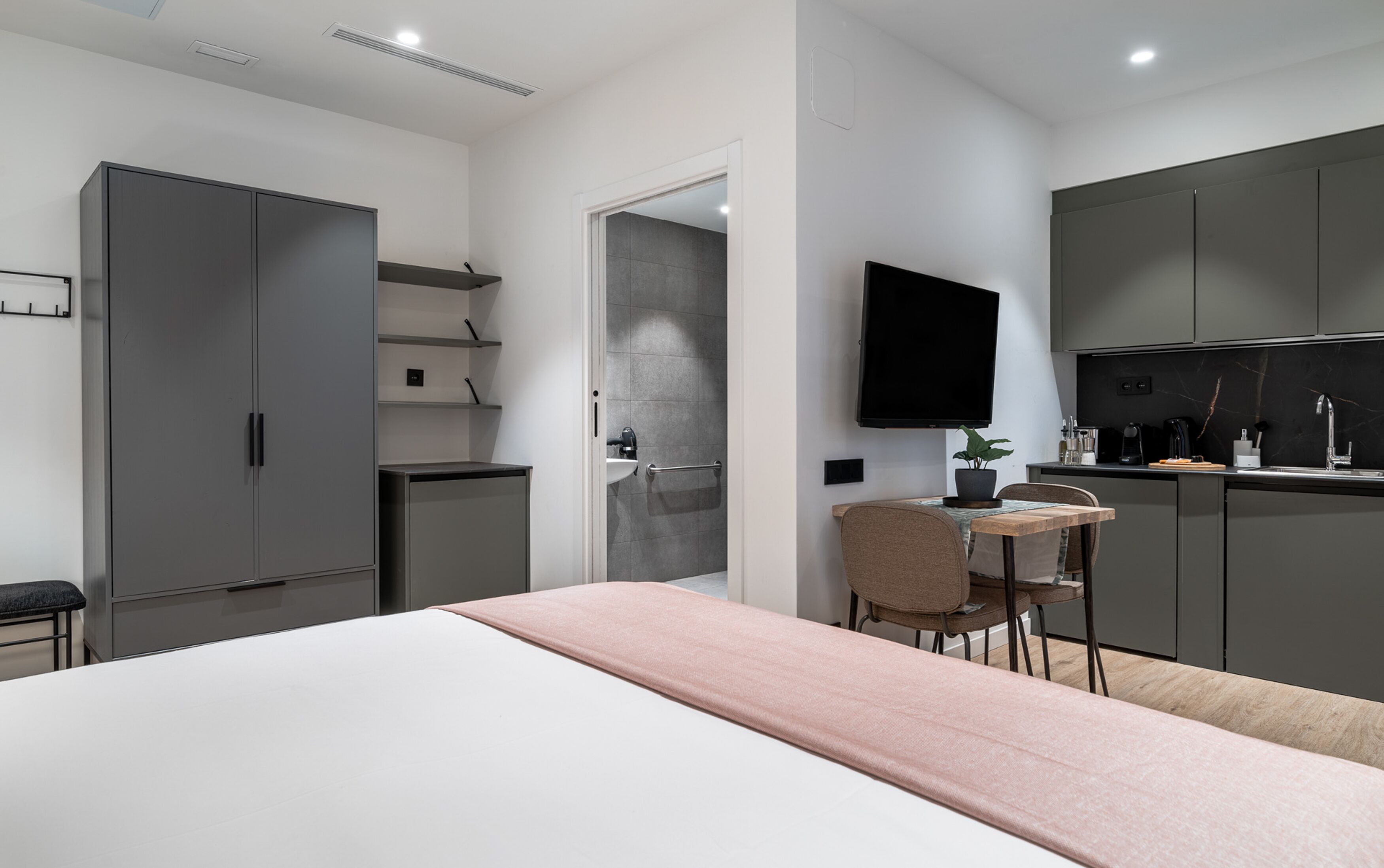 A modern bedroom with a bed, a bedside table, a chair, a TV, and a kitchenette.