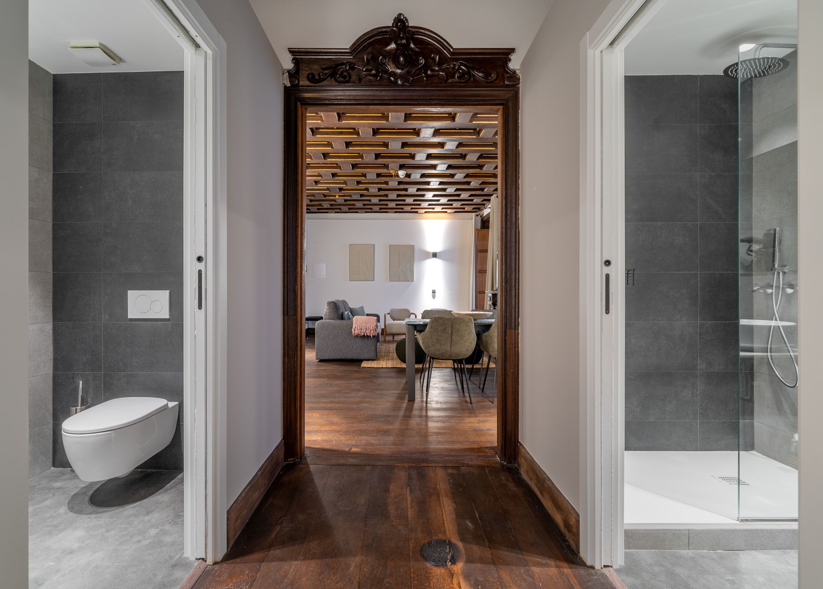 Un baño moderno con ducha, inodoro y una puerta de madera que da a una zona de estar con sofá y mesa de comedor.