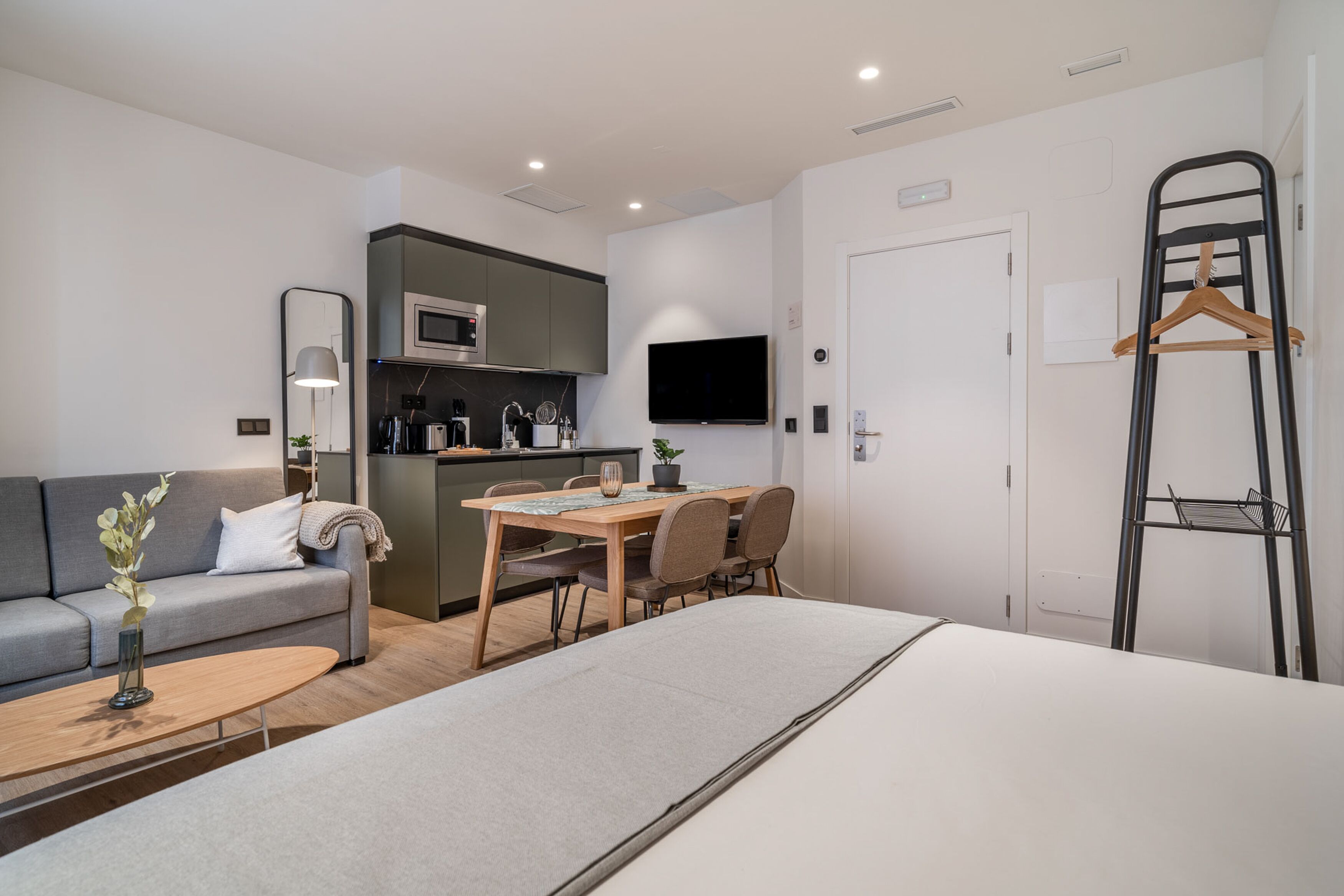 A modern hotel room with a bed, a sofa, a dining table, and a kitchenette.