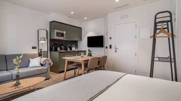 A modern hotel room with a bed, a sofa, a dining table, and a kitchenette.