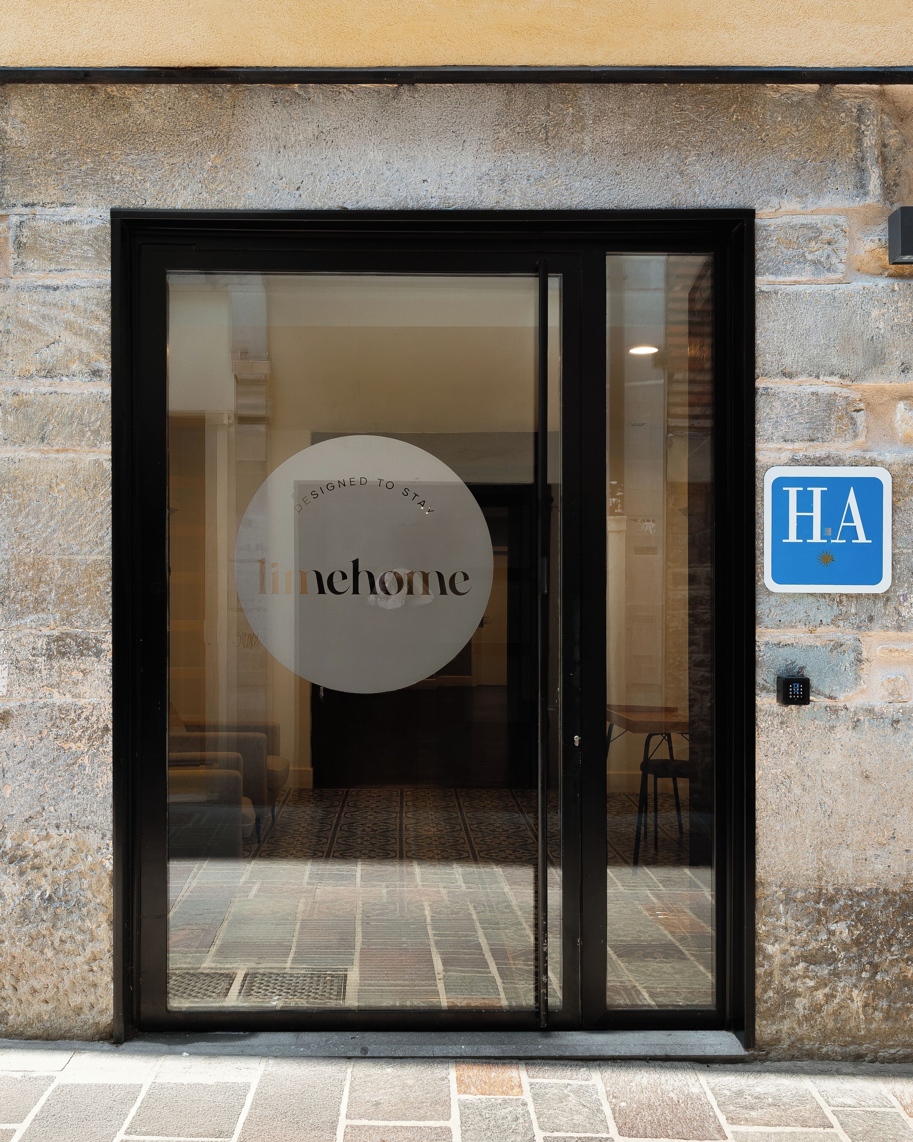 Ein Hotel Eingang mit einer Glastür, einem Schild mit der Aufschrift 'Designed to Stay at home' und einem blauen Schild mit 'HA' an der Wand.