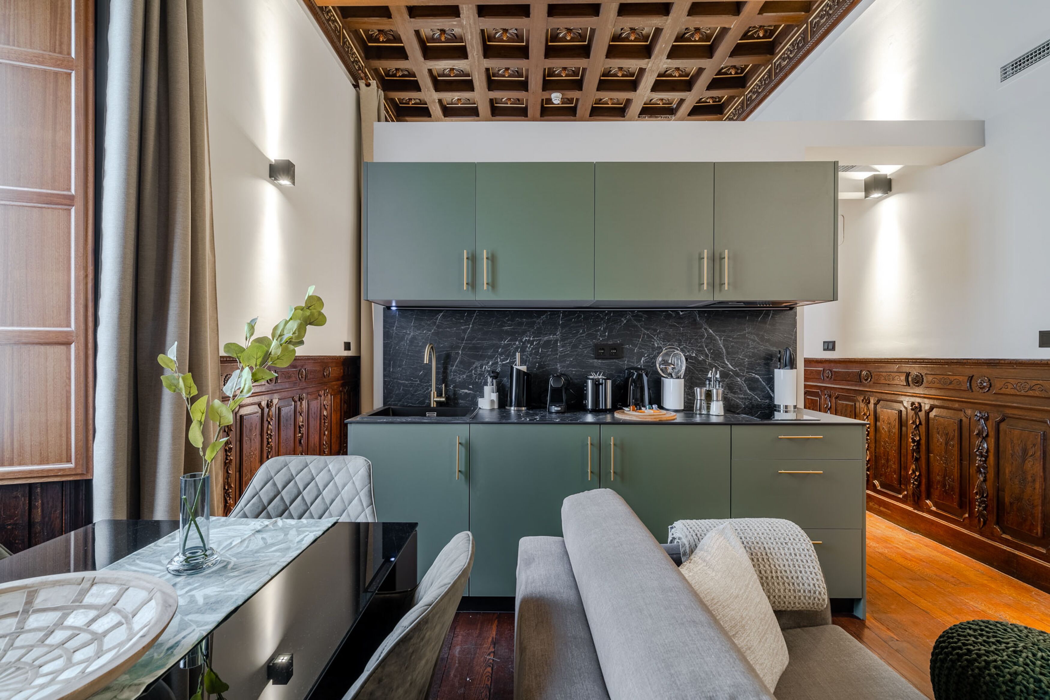 Una cocina moderna con armarios verdes, salpicadero de mármol y zona de comedor con mesa de vidrio y un jarrón de plantas verdes.