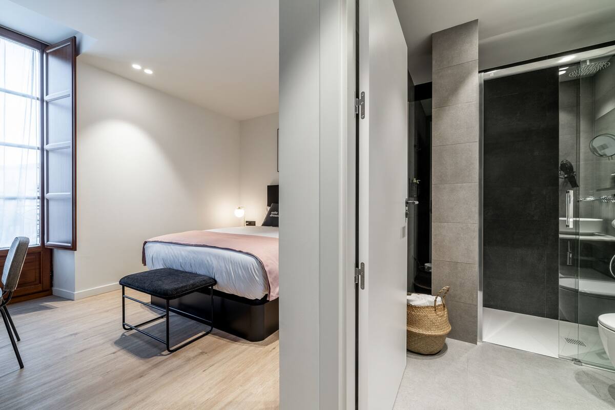 A modern bedroom with a bed, bedside table, and a chair. There is a bathroom with a shower, toilet, and a glass-enclosed shower area.