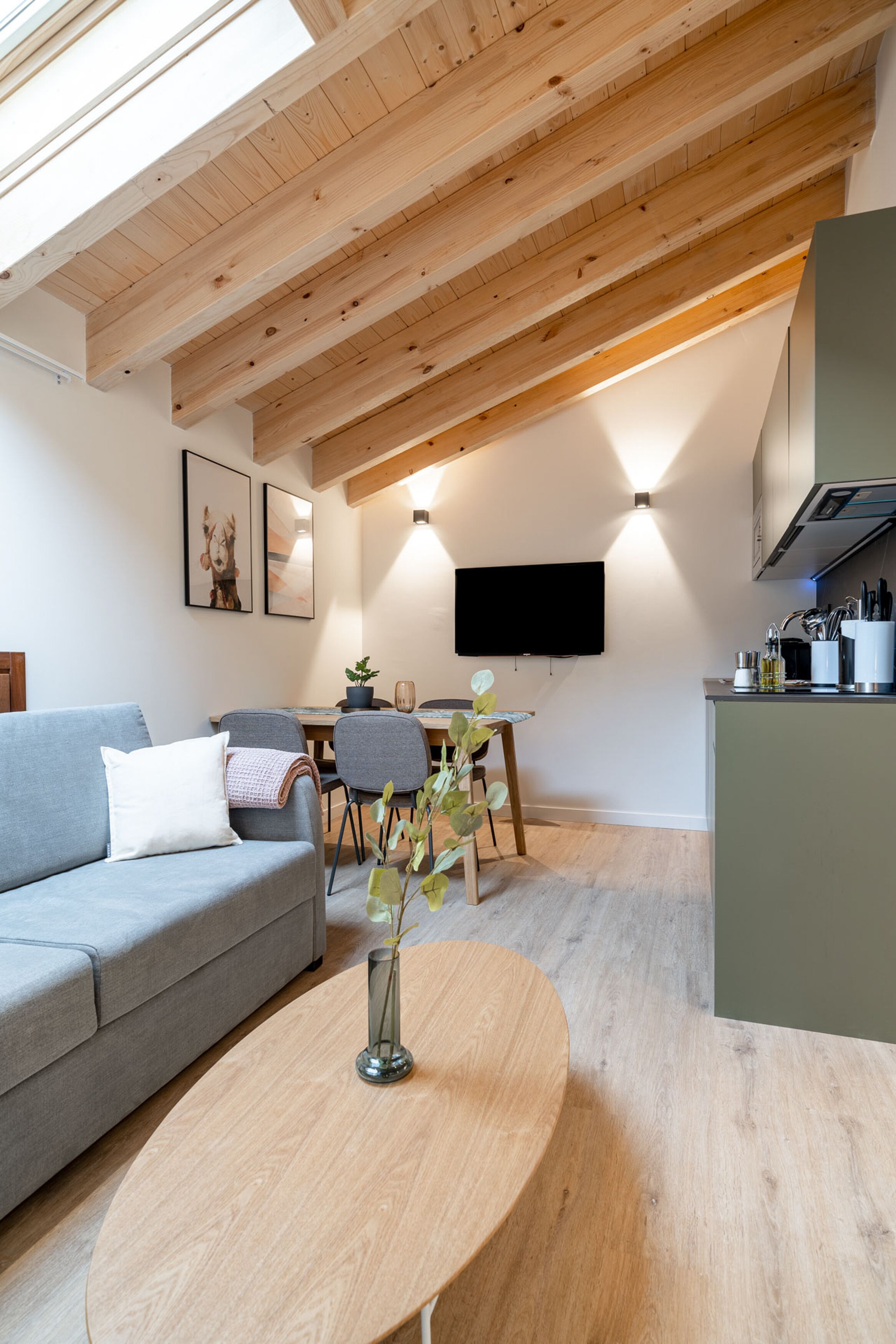 A modern living room with a grey sofa, a wooden coffee table, and a dining area with a TV and kitchen counter.