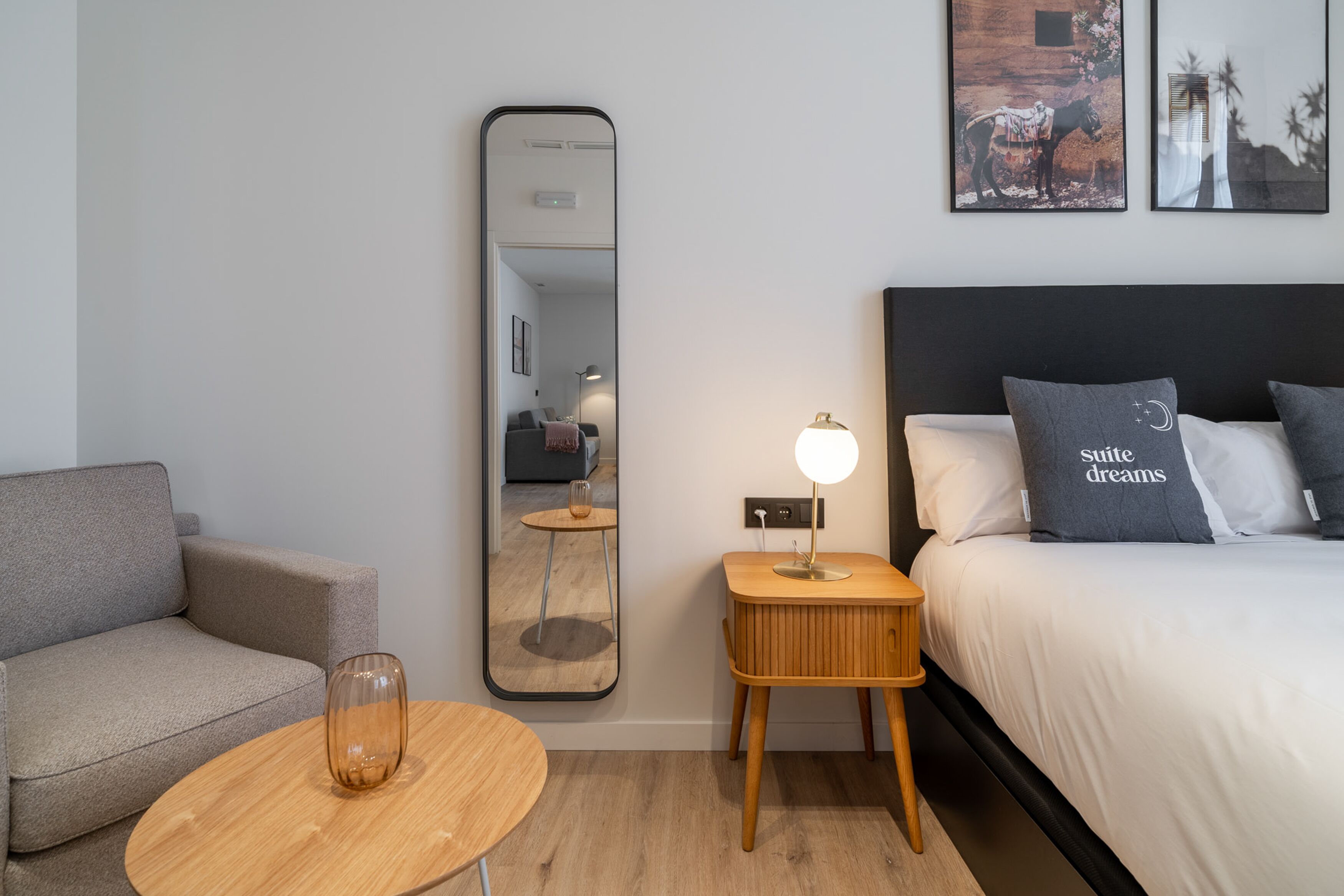 Een moderne hotelkamer met een bank, een ronde houten tafel, een bedlamp, een grote spiegel en een bed met twee kussens.