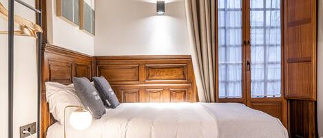 A bedroom with a wooden headboard, a bed with white bedding, a bedside table with a lamp, and a window with curtains.