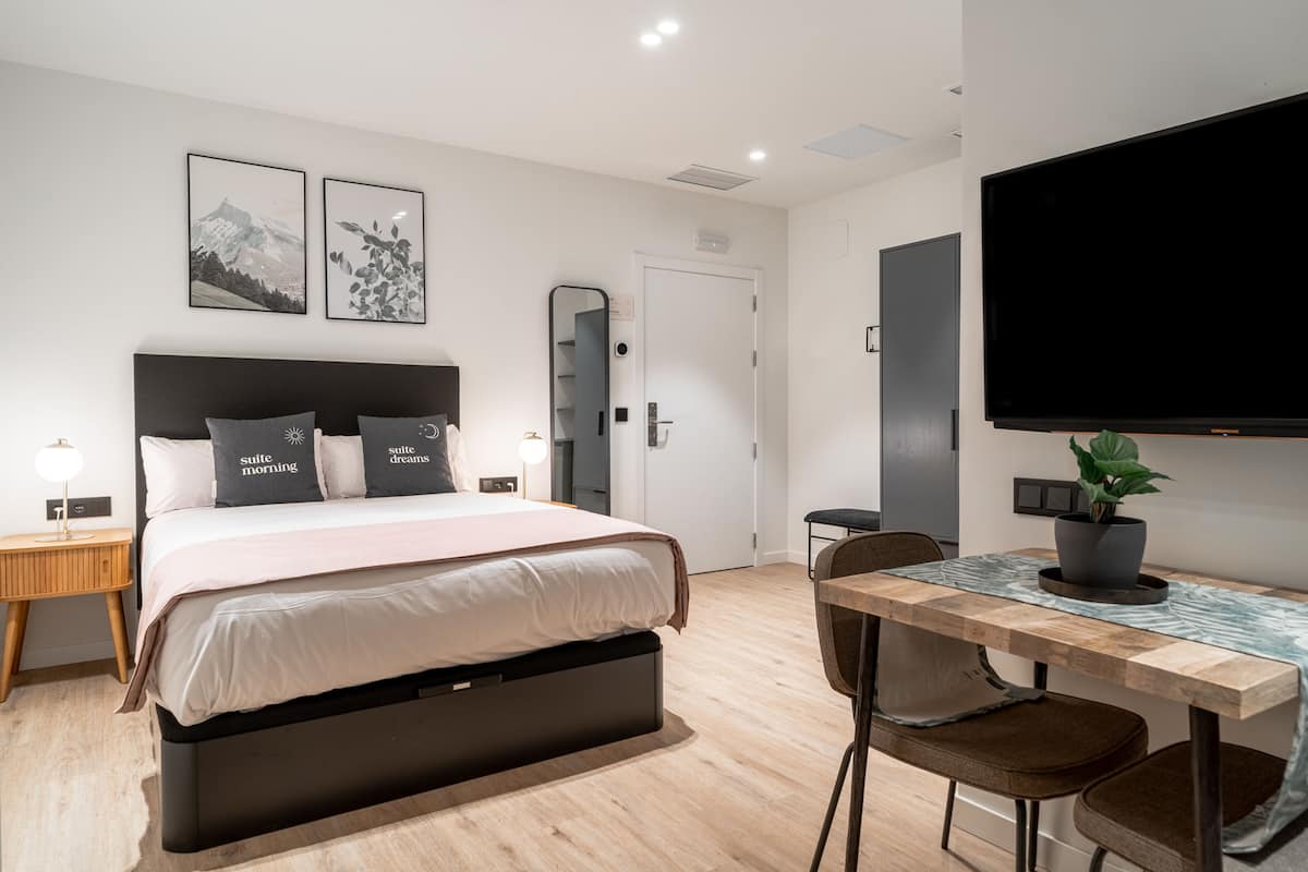 A modern hotel room with a bed, bedside table, chair, desk, and a flat-screen TV.