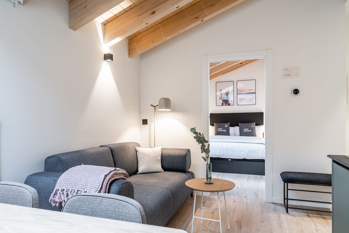 A modern bedroom with a grey sofa, a small round table, a bed, and a desk area.