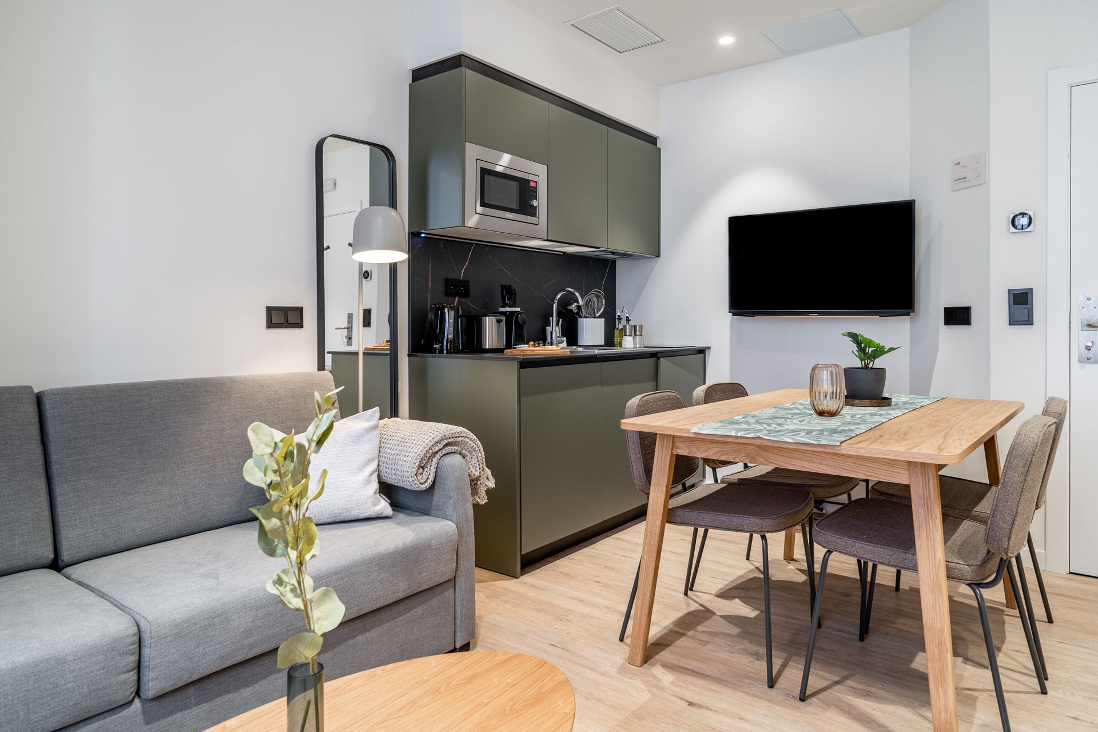 Una sala moderna con un sofá gris, una mesa de comedor de madera con cuatro sillas, una zona de cocina con microondas y un televisor de pantalla plana montado en la pared.
