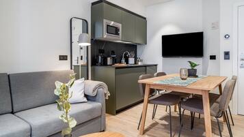 Una sala moderna con un sofá gris, una mesa de comedor de madera con cuatro sillas, una zona de cocina con microondas y un televisor de pantalla plana montado en la pared.