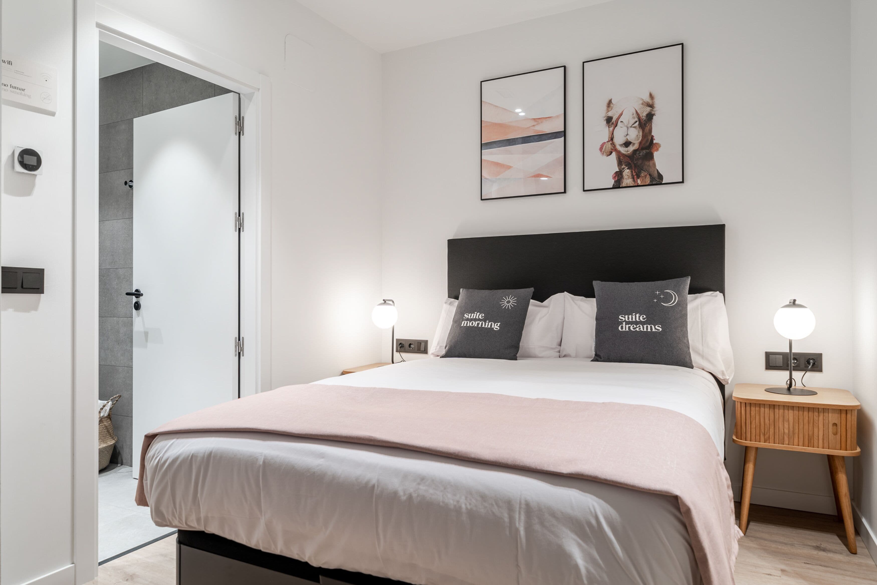 A modern bedroom with a large bed, two bedside lamps, a nightstand, and a door leading to a bathroom.