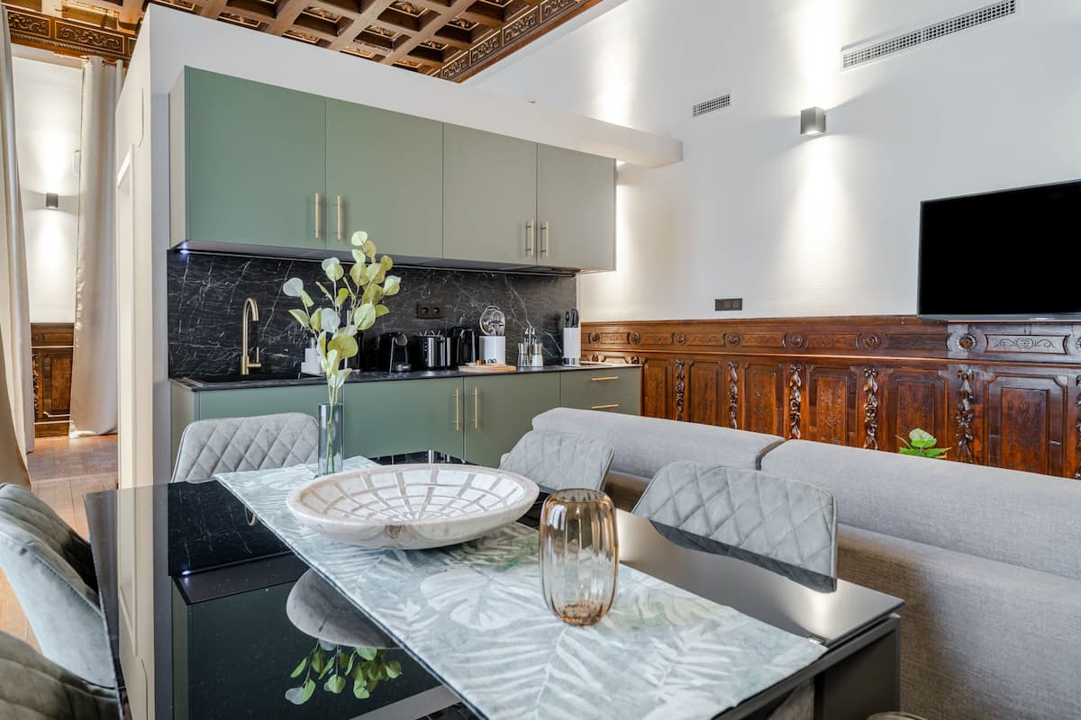 A modern kitchen with green cabinets, a marble countertop, and a dining area with a glass table and grey upholstered sofa.
