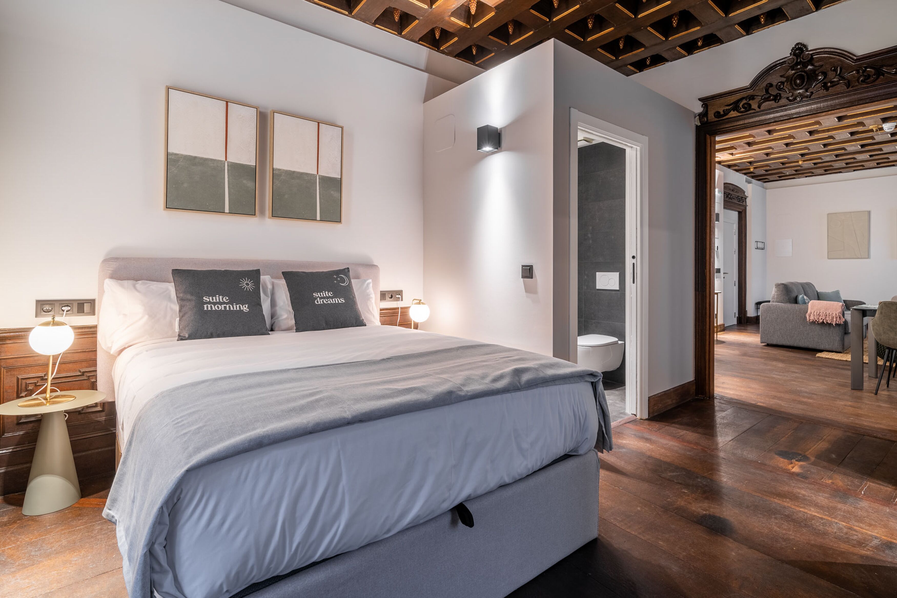Un dormitorio moderno con una cama grande, mesitas de noche y una mesita auxiliar. Hay una puerta que conduce a otra habitación.