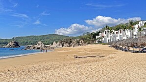 A beach with white buildings, palm trees, and a clear blue sky. - Beachfront condo with ocean view, private pool & resort amenities (Bahías de Huatulco)