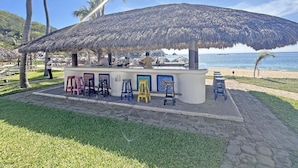 A beachside bar with a thatched roof, outdoor seating, and a view of the ocean. - Beachfront condo with ocean view, private pool & resort amenities (Bahías de Huatulco)