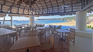An outdoor seating area with tables and chairs, a thatched roof, and a view of the beach and ocean. - Beachfront condo with ocean view, private pool & resort amenities (Bahías de Huatulco)