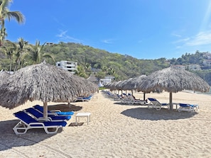 A beach with rows of sun loungers under thatched umbrellas, a sandy beach, and a backdrop of hills and buildings. - Beachfront condo with ocean view, private pool & resort amenities (Bahías de Huatulco)