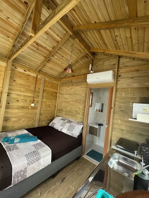 A wooden cabin room with a bed, a hanging light fixture, an air conditioning unit, a small bathroom with a sink, and a stove. - Floripa Glamping (Florianópolis)