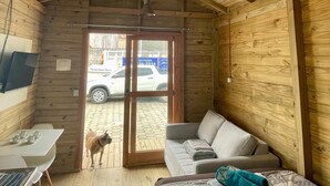 Une cabane chaleureuse aux murs en bois et au plafond incliné. À l’intérieur, on trouve un lit, un canapé et une table avec des tasses. On peut apercevoir un chien à travers la porte.