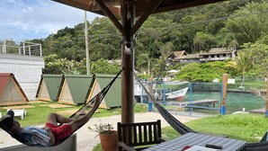 Un hamac sous une pergola en bois, une table à carreaux avec un ordinateur portable et un verre, et une vue sur une étendue d’eau et des bâtiments au loin.