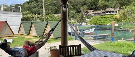 Una hamaca bajo una pérgola de madera, una mesa a cuadros con una computadora portátil y un vaso, y una vista de un cuerpo de agua y edificios al fondo.