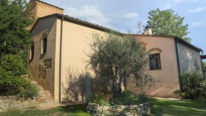 A two-story building with a stone staircase leading to the entrance, surrounded by a well-maintained garden with various plants and flowers. - Cammino Santo - studio w/private pool & garden (Poggibonsi)