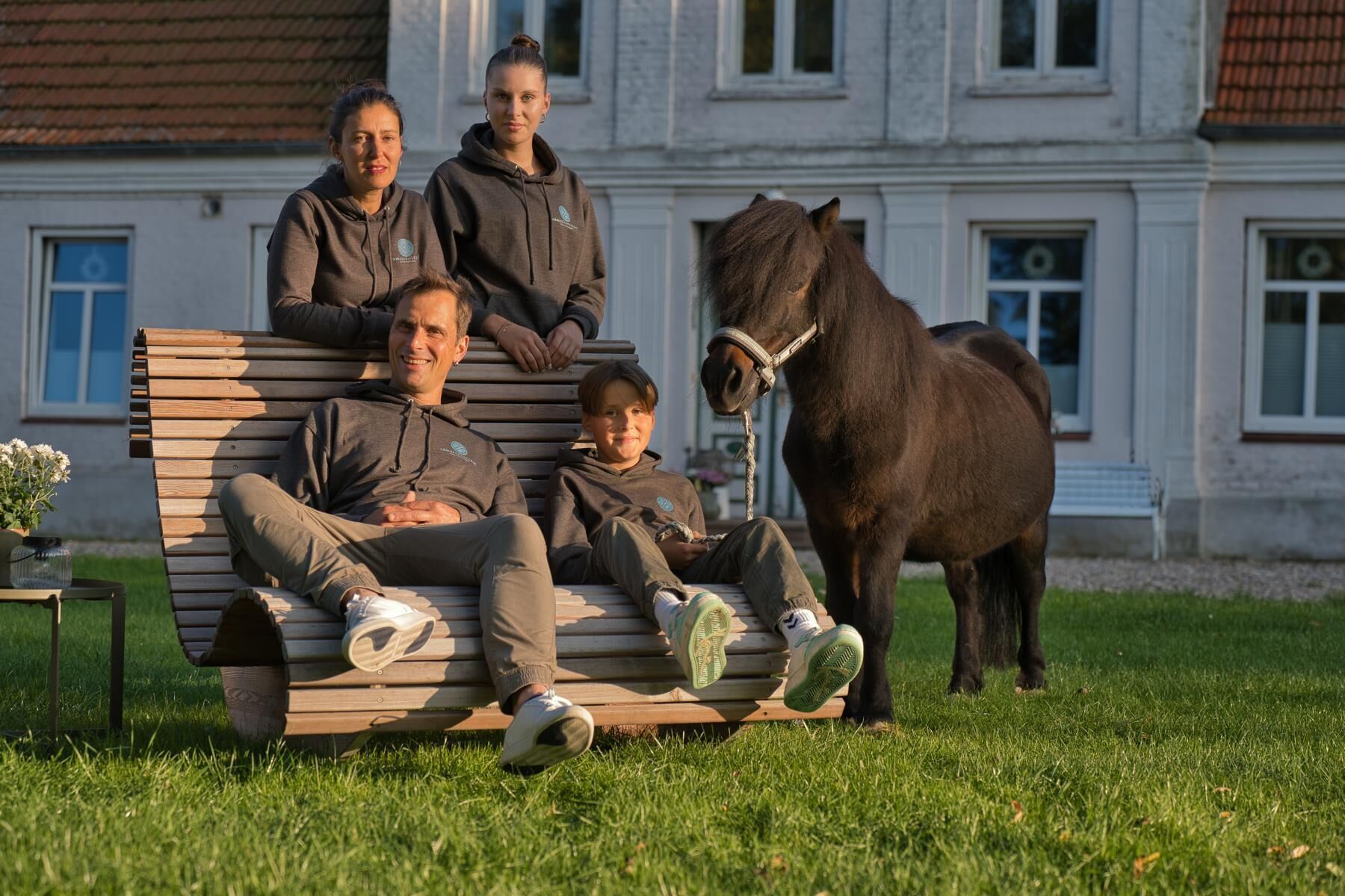 A family sitting on a wooden bench outdoors with a horse nearby.