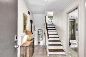 A modern hallway with a wooden staircase, a door with a security camera, and a view into a bathroom.