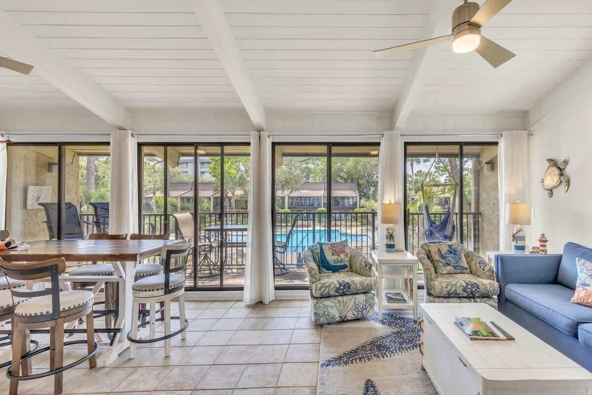 A spacious living area with a dining table, chairs, a sofa, and a coffee table. There is a ceiling fan, large windows, and a view of a swimming pool.