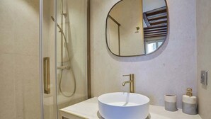 Een moderne badkamer met een douche met glazen wand, een ronde spiegel en een wit wastafelmeubel met een gouden kraan.